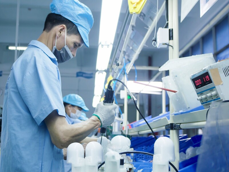 Worker assembling electronic parts at a lit workstation on the production floor.